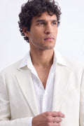 A man with curly dark hair wearing a light cream-colored hemp-blend blazer over an open white dress shirt, photographed against a clean white studio background.