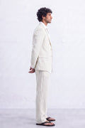 A man in a side profile wearing a light cream-colored hemp suit, white dress shirt, and black sandals, standing against a textured white wall.
