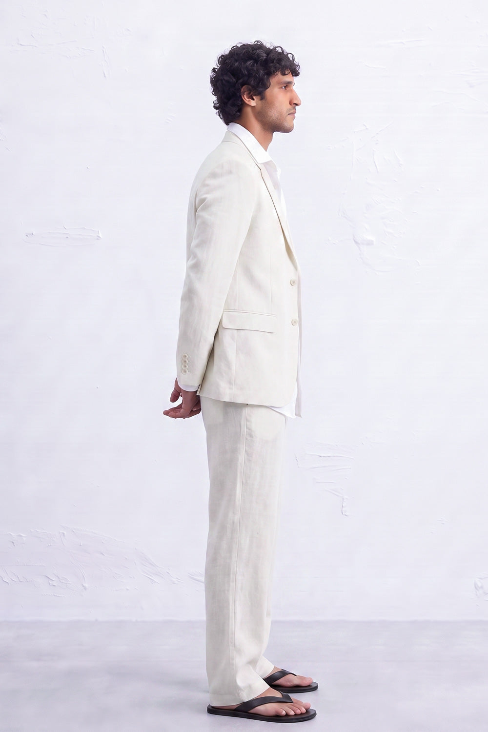 A man in a side profile wearing a light cream-colored hemp suit, white dress shirt, and black sandals, standing against a textured white wall.