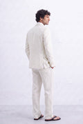 A man wearing a light beige hemp two-piece suit, consisting of a blazer and trousers, paired with dark brown sandals, photographed from behind against a textured white wall.