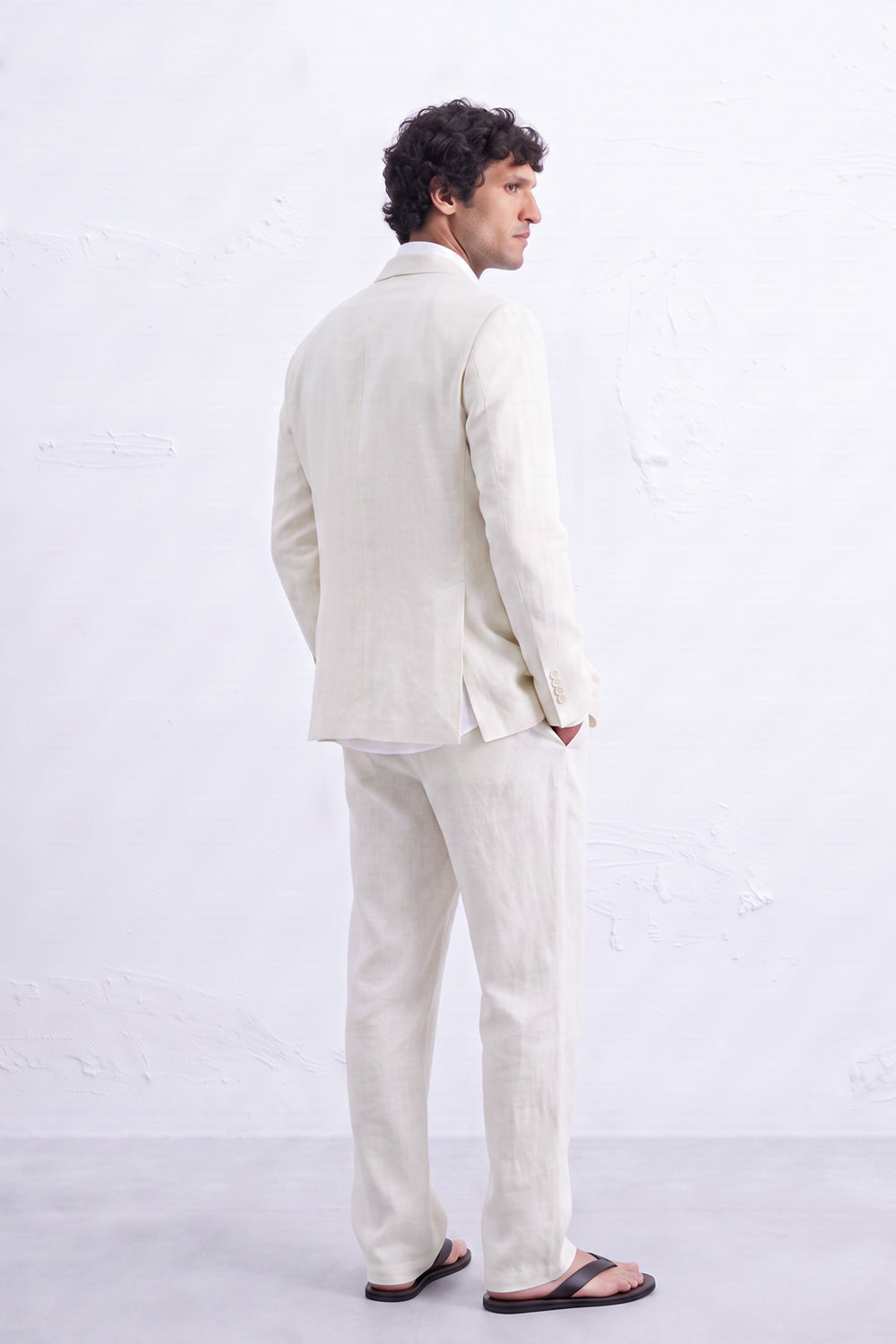 A man wearing a light beige hemp two-piece suit, consisting of a blazer and trousers, paired with dark brown sandals, photographed from behind against a textured white wall.