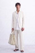 A man modeling a light cream hemp suit consisting of a blazer and trousers, paired with a white shirt, black sandals, and holding a matching cream canvas tote bag against a white brick wall background.