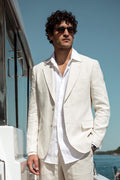 A man wearing a light cream hemp suit jacket and trousers with a white dress shirt, standing on a yacht deck with blue water and sky in the background.