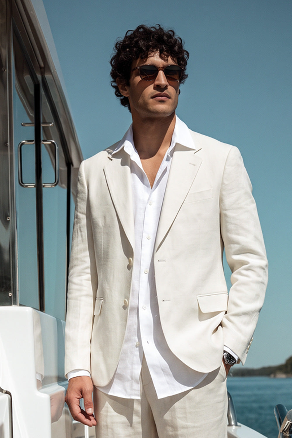 A man wearing a light cream hemp suit jacket and trousers with a white dress shirt, standing on a yacht deck with blue water and sky in the background.