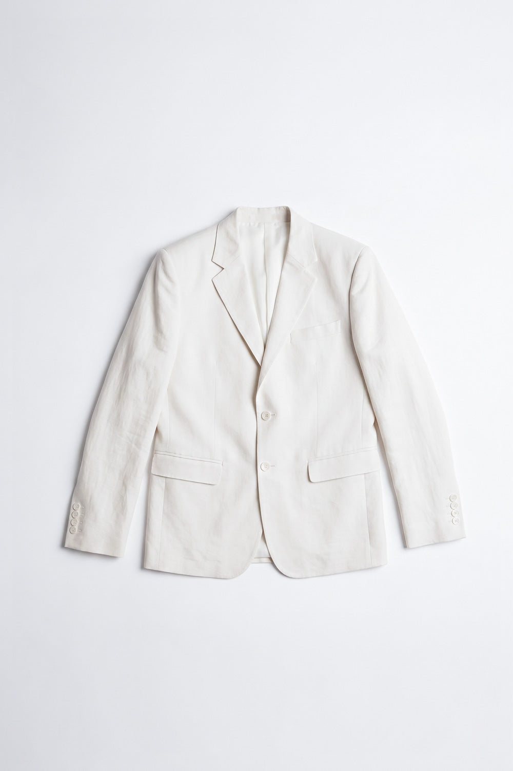 A man modeling a light cream hemp suit consisting of a blazer and trousers, worn over a white shirt and black sandals, standing against a white textured wall.