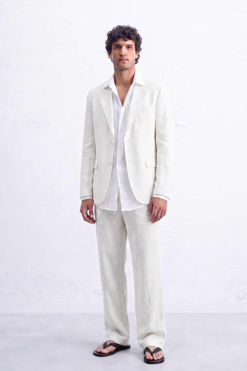 A man modeling a light cream hemp suit consisting of a blazer and trousers, worn over a white shirt and black sandals, standing against a white textured wall.