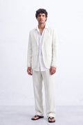 A man modeling a light cream hemp suit consisting of a blazer and trousers, worn over a white shirt and black sandals, standing against a white textured wall.