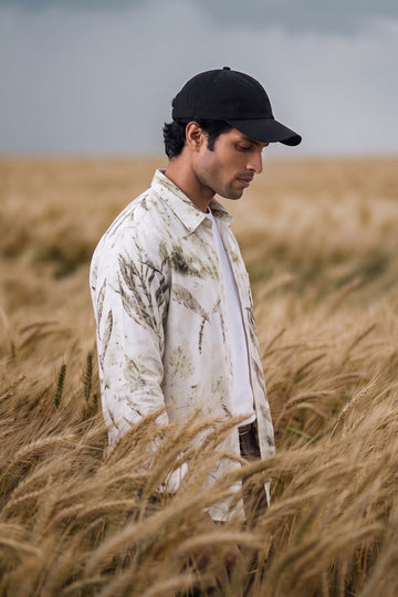 A man standing in a golden wheat field, wearing a black baseball cap, a white button-up shirt with a brown leaf pattern, and a white undershirt.
