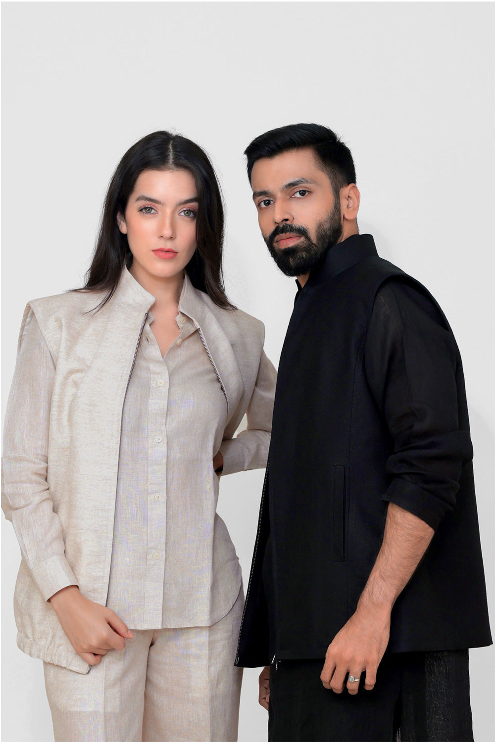 A man and a woman modeling coordinated apparel against a plain light gray studio background. The woman wears a light beige tailored slinen vest with a collared shirt, and the man wears a black sleeveless linen vest over a black long-sleeve top.