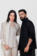 A man and a woman modeling coordinated apparel against a plain light gray studio background. The woman wears a light beige tailored slinen vest with a collared shirt, and the man wears a black sleeveless linen vest over a black long-sleeve top.