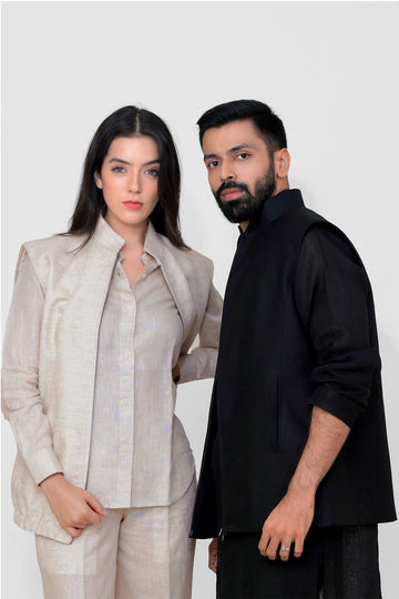 A man and a woman modeling coordinated apparel against a plain light gray studio background. The woman wears a light beige tailored slinen vest with a collared shirt, and the man wears a black sleeveless linen vest over a black long-sleeve top.