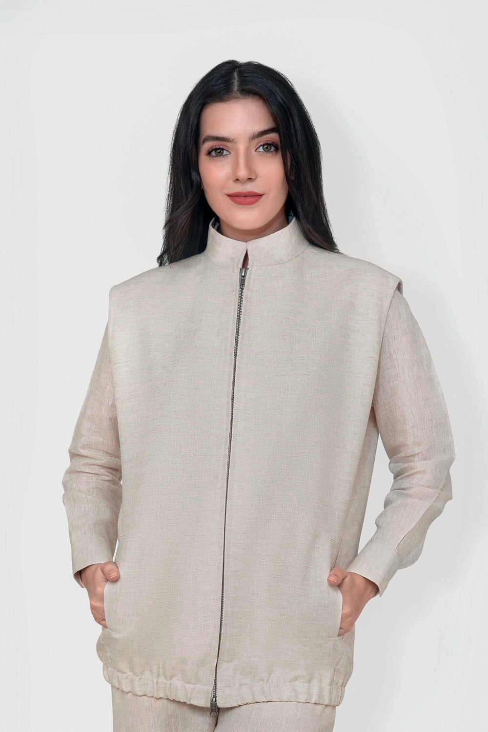 A woman modeling a light beige sleeveless zip-up linen vest with a high stand-up collar and front pockets, set against a plain light gray studio background.