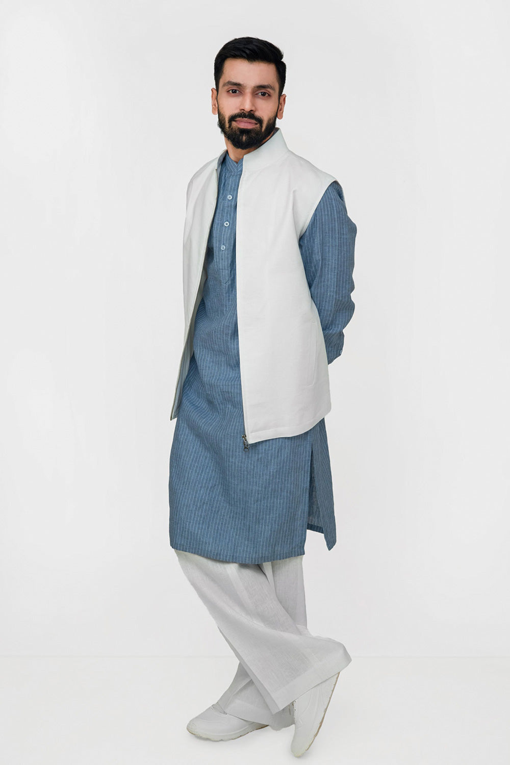 A man modeling a traditional South Asian outfit consisting of a light blue textured kurta, a white sleeveless linen vest, and light gray trousers, set against a plain white studio background.
