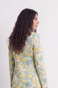 A woman with long, wavy dark brown hair is shown from a side-back angle, wearing a long-sleeve silk blazer with a watercolor-style botanical leaf pattern in yellow, sage green, and cream.