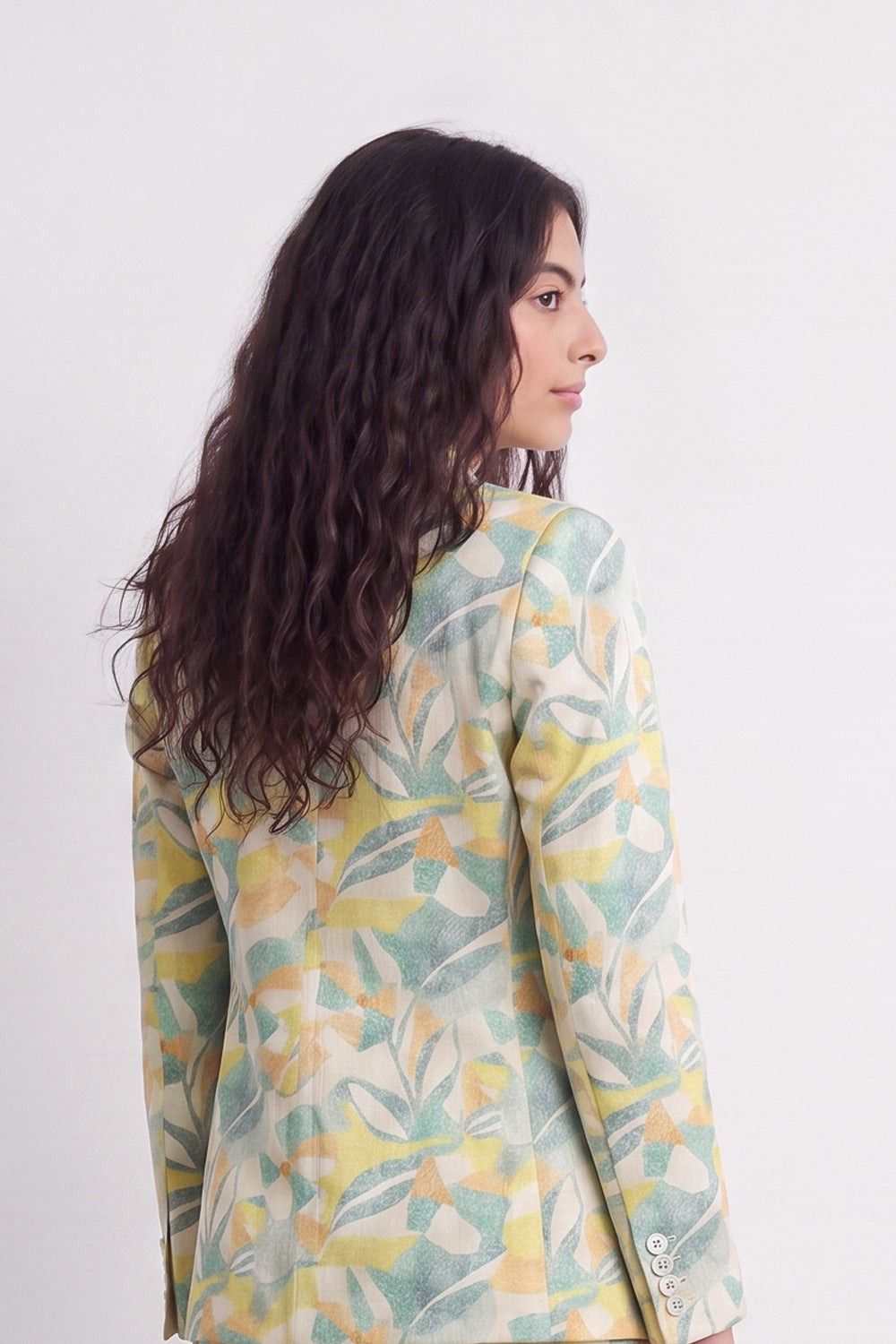 A woman with long, wavy dark brown hair is shown from a side-back angle, wearing a long-sleeve silk blazer with a watercolor-style botanical leaf pattern in yellow, sage green, and cream.