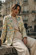A woman sits on a stone ledge in front of a European-style building, wearing a light beige outfit with a yellow, green, and blue floral patterned silk blazer.