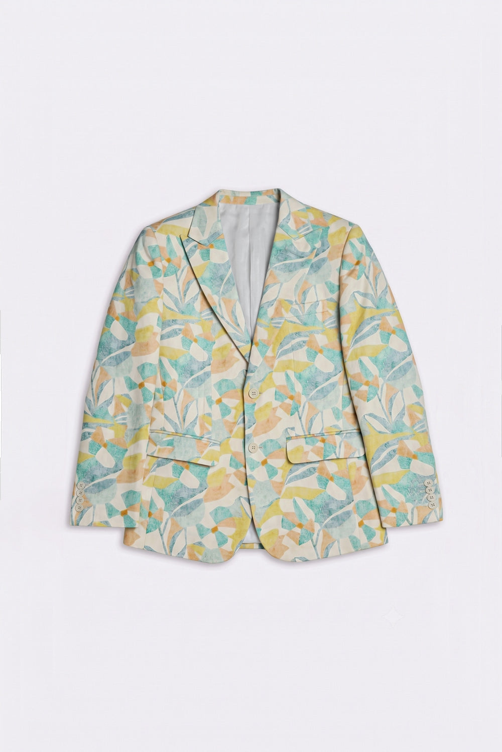 A woman stands against a plain light gray studio wall wearing a beige satin-like blouse and wide-leg beige trousers, with a patterned silk blazer draped over her shoulders. The blazer has a yellow and mint green abstract floral or leaf print.
