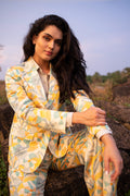 A woman wearing a matching yellow, green, and white patterned silk blazer and trousers set, sitting outdoors on a rock against a soft sky background.