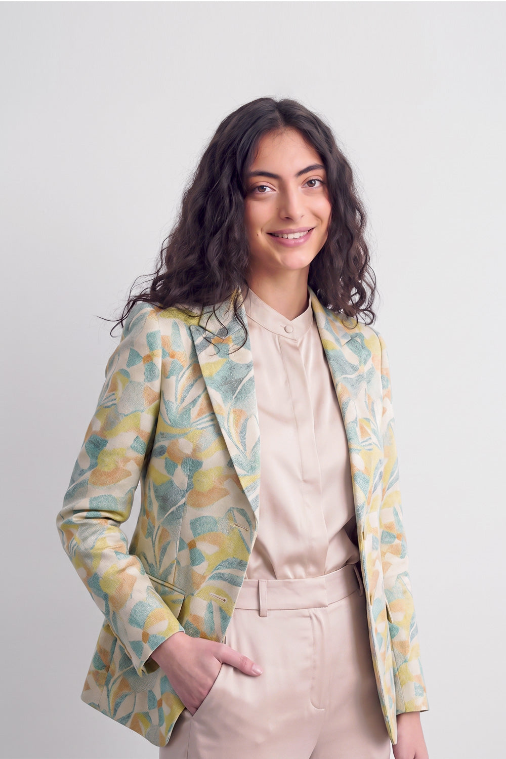 A woman modeling a light beige blouse and matching trousers, styled with a patterned silk blazer featuring abstract yellow, green, and blue floral motifs, against a plain light gray background.