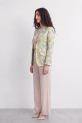 A woman stands in profile wearing a light beige top and wide-leg beige trousers, with a pastel tropical print silk blazer in yellow, green, and blue over her shoulders, against a plain white studio background.