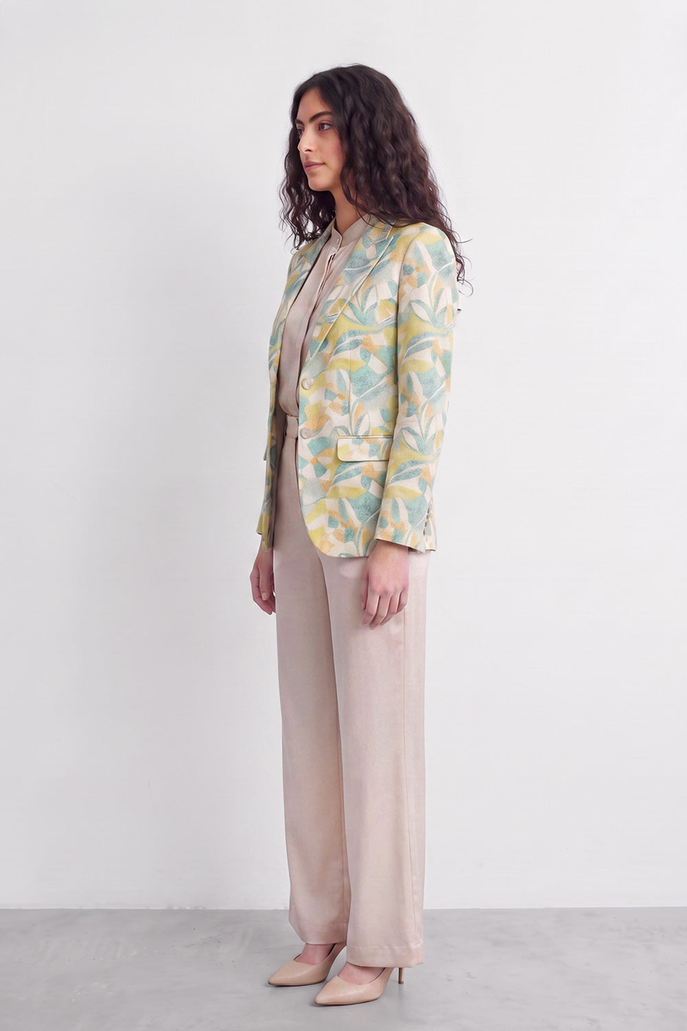 A woman stands in profile wearing a light beige top and wide-leg beige trousers, with a pastel tropical print silk blazer in yellow, green, and blue over her shoulders, against a plain white studio background.