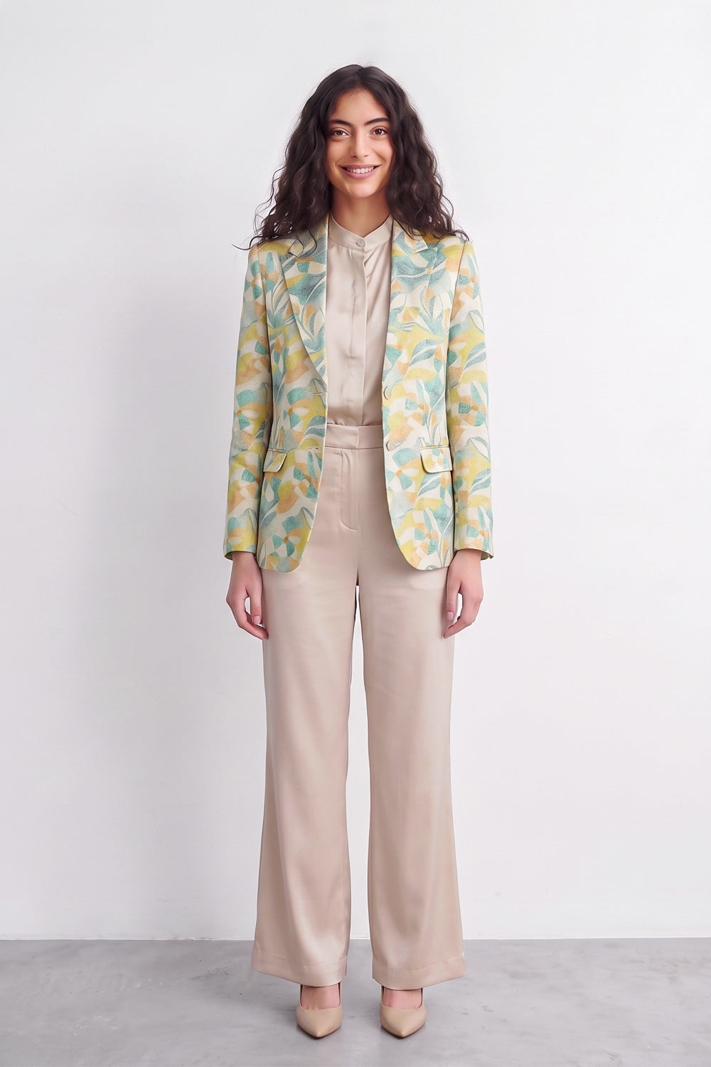 A woman stands against a plain light gray studio wall wearing a beige satin-like blouse and wide-leg beige trousers, with a patterned silk blazer draped over her shoulders. The blazer has a yellow and mint green abstract floral or leaf print.