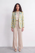 A woman stands against a plain light gray studio wall wearing a beige satin-like blouse and wide-leg beige trousers, with a patterned silk blazer draped over her shoulders. The blazer has a yellow and mint green abstract floral or leaf print.