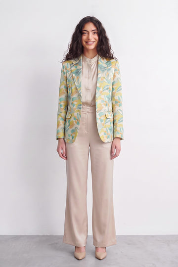 A woman stands against a plain light gray studio wall wearing a beige satin-like blouse and wide-leg beige trousers, with a patterned silk blazer draped over her shoulders. The blazer has a yellow and mint green abstract floral or leaf print.