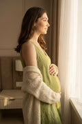 A pregnant woman in profile stands by a window wearing a sage green maternity hemp dress and a cream knit cardigan, with warm sunlight streaming in from the right.