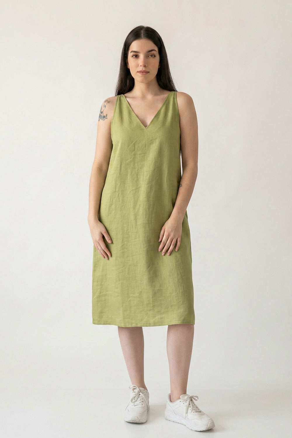 A woman modeling a sleeveless olive green hemp midi dress with a V-neckline, paired with white sneakers, against a plain off-white studio background.