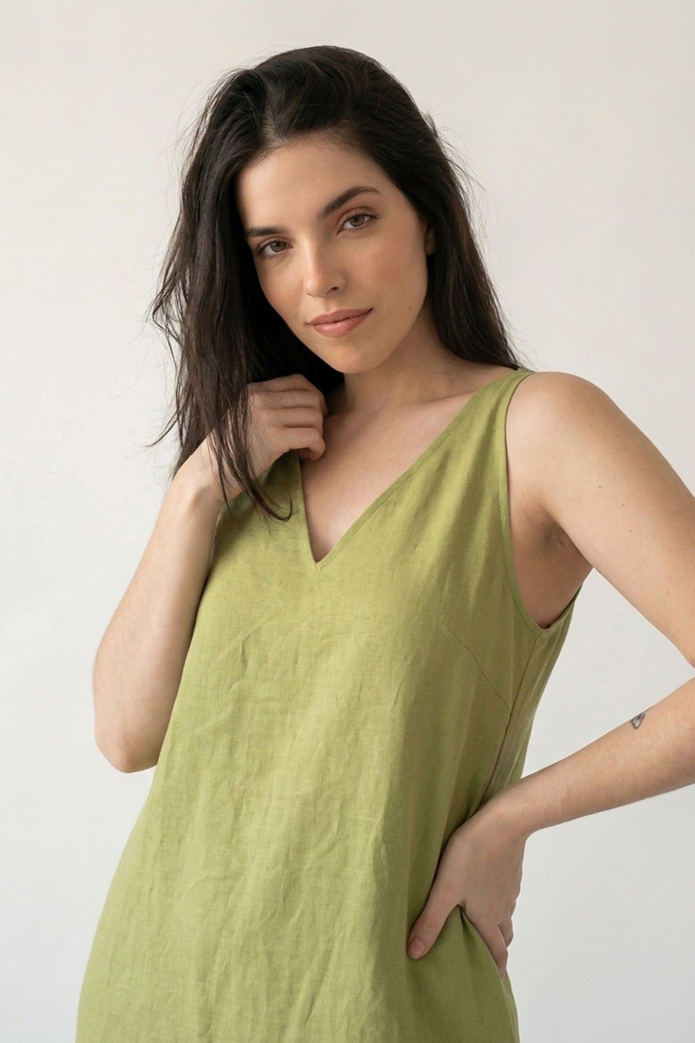 A woman modeling a sleeveless olive green V-neck hemp dress against a plain off-white background.