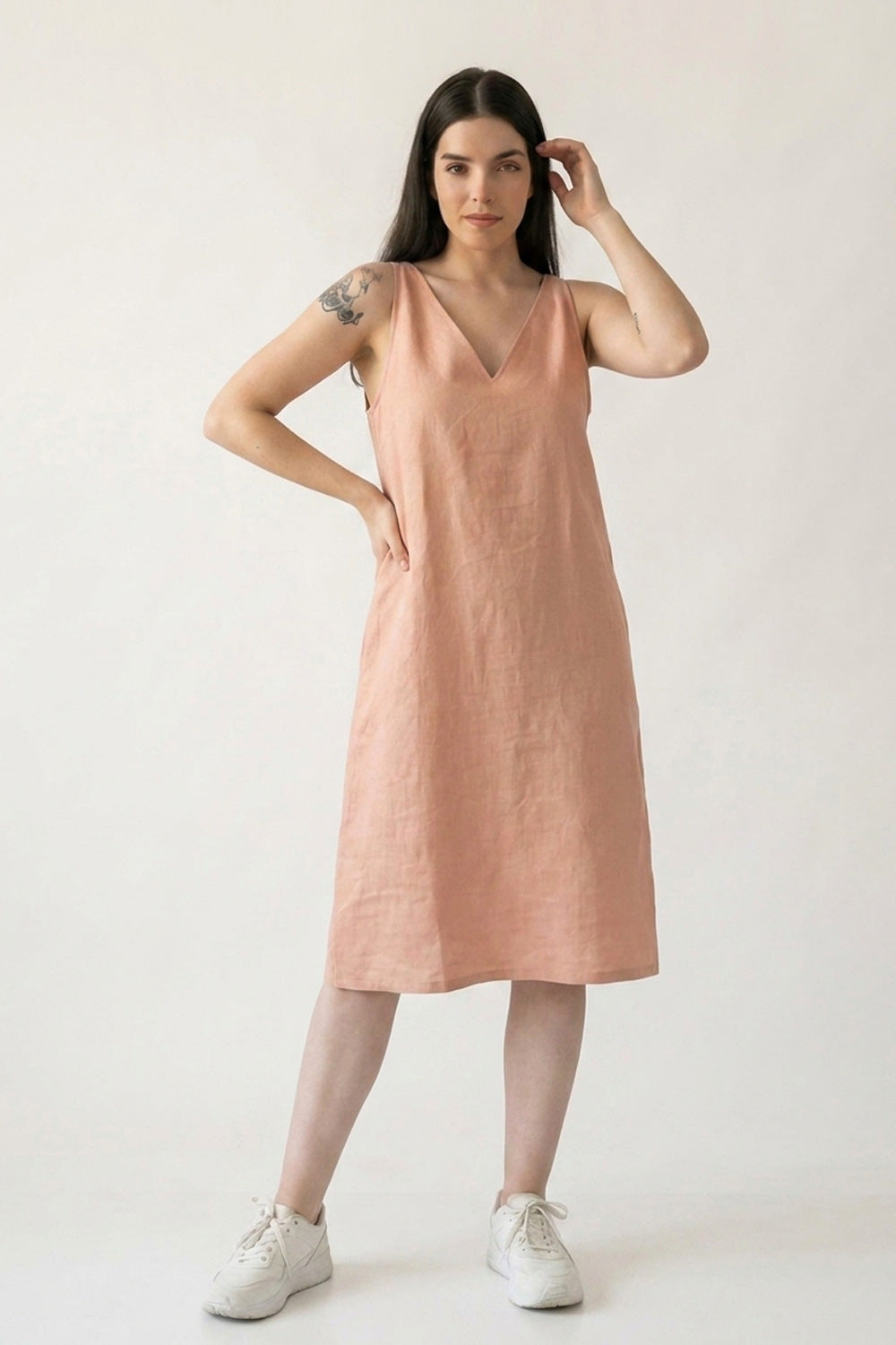 A woman modeling a sleeveless, V-neck midi hemp dress in a dusty peach-pink color, paired with white sneakers, against a plain light gray studio background.