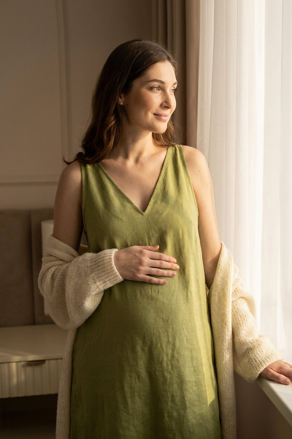 A pregnant woman wearing a sage green hemp slip dress and a cream knit cardigan, standing by a window in a softly lit room.