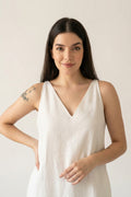 A woman with long dark hair wearing a sleeveless white V-neck hemp dress, standing against a plain light beige background.