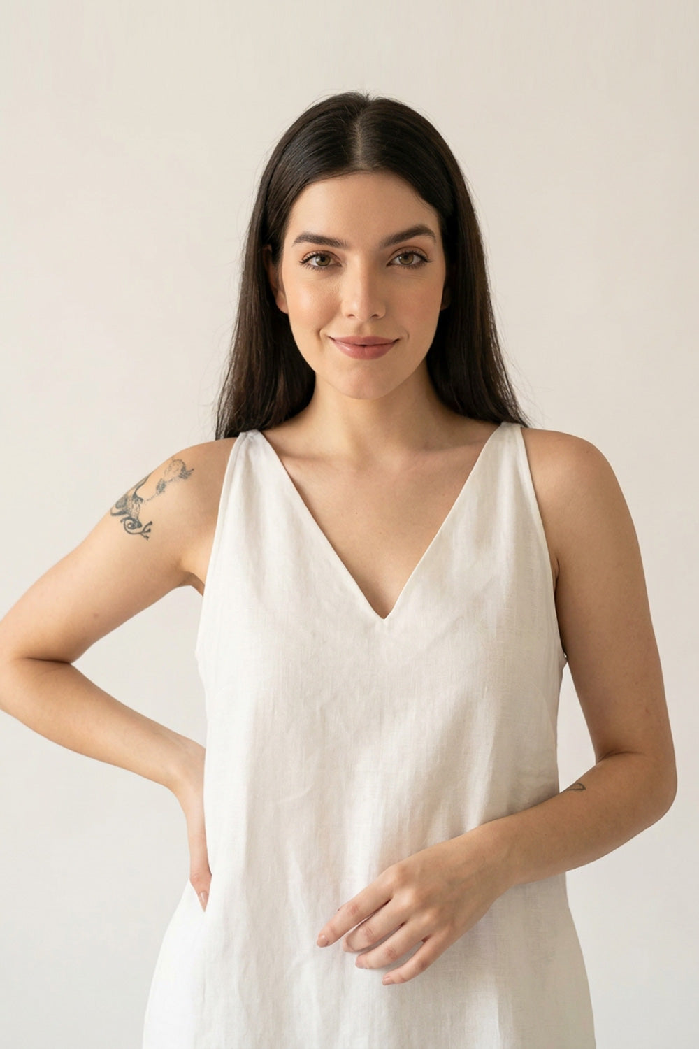 A woman with long dark hair wearing a sleeveless white V-neck hemp dress, standing against a plain light beige background.