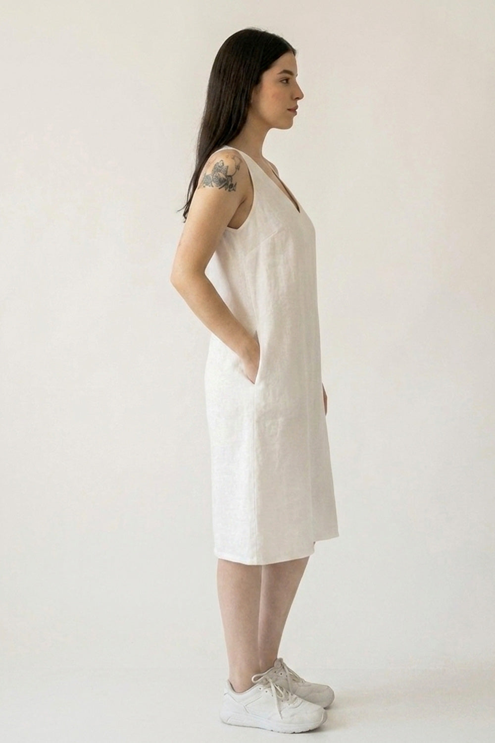 A woman in profile wearing a sleeveless white hemp midi dress with side pockets, paired with white sneakers, standing against a plain off-white studio background.