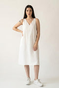 A woman modeling a sleeveless white hemp midi dress with a V-neckline and side pockets, paired with white sneakers, against a plain off-white studio background.