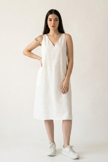 A woman modeling a sleeveless white hemp midi dress with a V-neckline and side pockets, paired with white sneakers, against a plain off-white studio background.