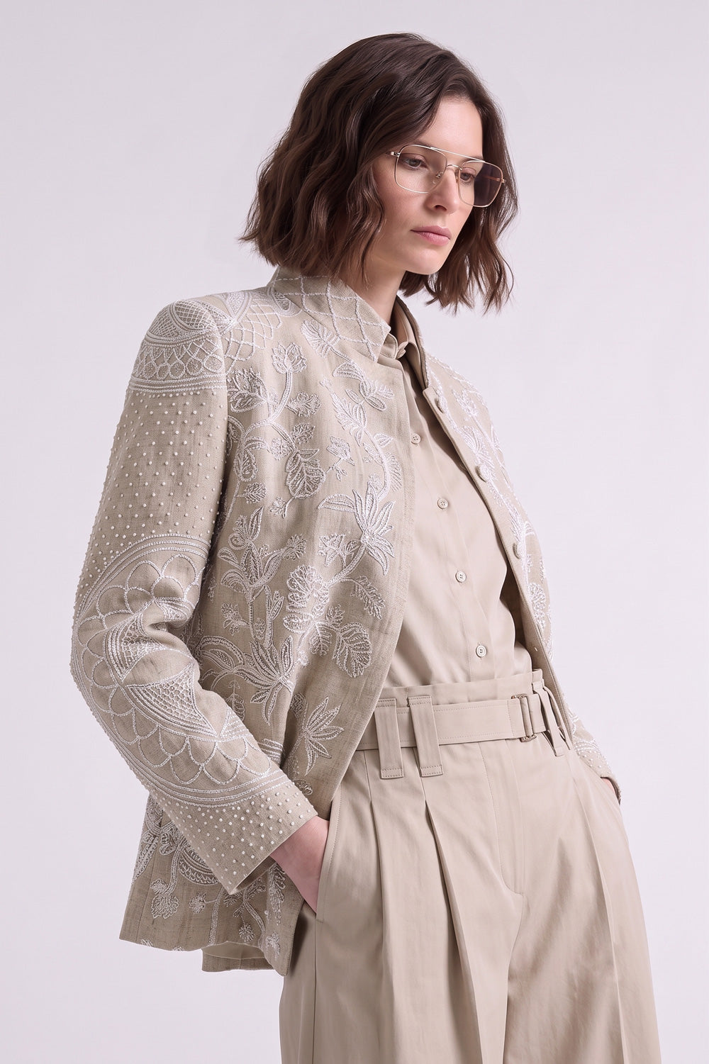 A woman wearing a beige embroidered blazer over a matching beige button-up shirt and pleated trousers, photographed against a clean light gray studio background.