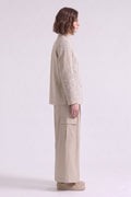 A model shown in profile wearing a beige cropped linen jacket with intricate white embroidery on the sleeves and matching beige wide-leg cargo pants, set against a plain light gray studio background.