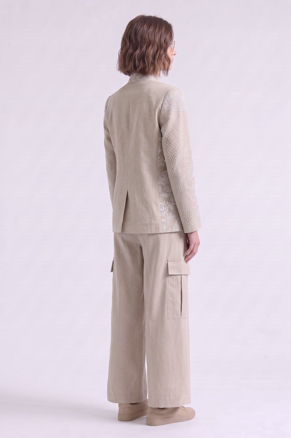 A model shown from the back wearing a light beige textured linen jacket and matching wide-leg cargo pants against a plain light gray studio background.