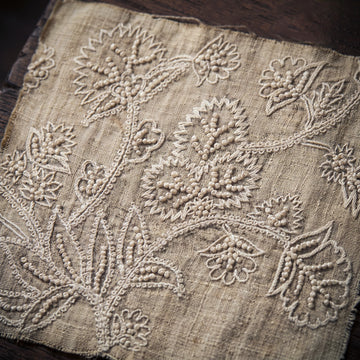 A close-up of a beige linen fabric featuring intricate floral embroidery with raised beadwork, laid flat on a dark wooden surface.