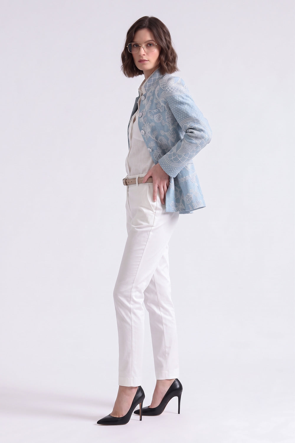 A woman modeling a light blue embroidered linen blazer, white top and trousers, and black heels against a plain white studio background.