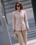 A woman wearing a beige and white embroidered linen jacket with matching beige trousers, styled with sunglasses and holding a beige clutch bag, set against an urban street background.