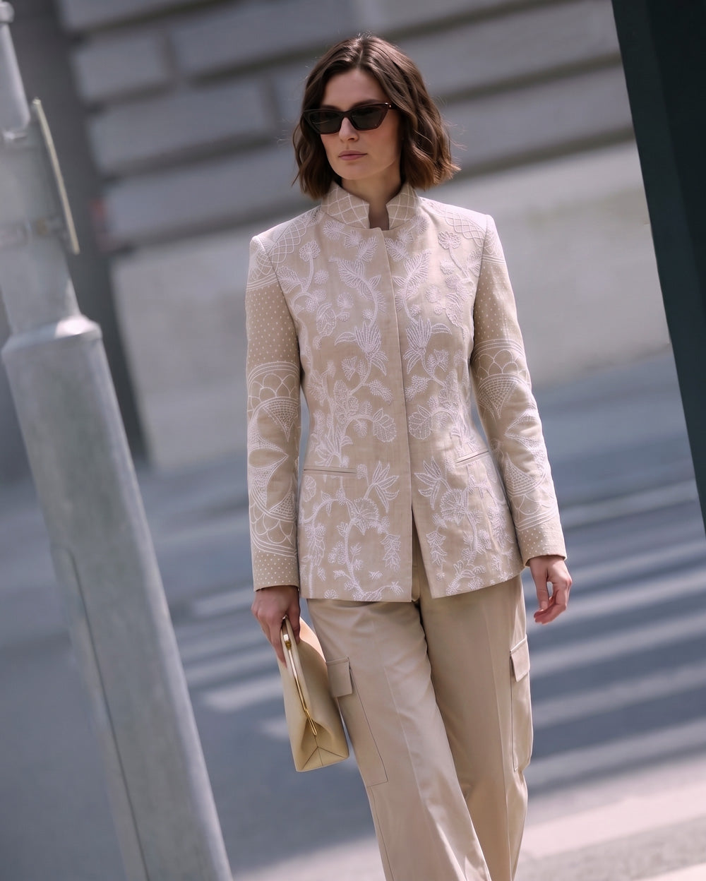 A woman wearing a beige and white embroidered linen jacket with matching beige trousers, styled with sunglasses and holding a beige clutch bag, set against an urban street background.