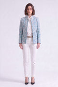 A woman stands against a light gray studio background wearing a light linen blue embroidered blazer over a white collared shirt, white pants, and black pointed heels.