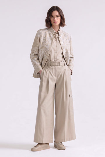 A model stands against a light gray studio background wearing a beige and cream embroidred linen jacket over a matching beige shirt and wide-leg trousers, paired with beige sneakers.