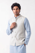 A man wearing a light grey tailored linen waistcoat over a pale blue long-sleeve shirt, standing against a neutral studio background.