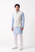 A full-length studio shot of a man wearing a light grey tailored waistcoat over a pale blue kurta, paired with white trousers and white sneakers against a neutral background.