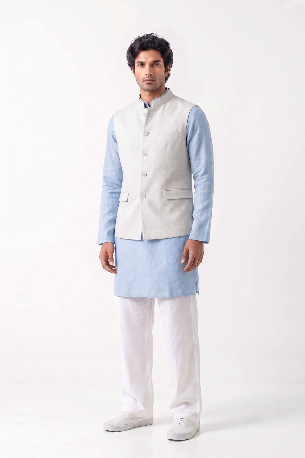 A full-length studio shot of a man wearing a light grey tailored waistcoat over a pale blue kurta, paired with white trousers and white sneakers against a neutral background.